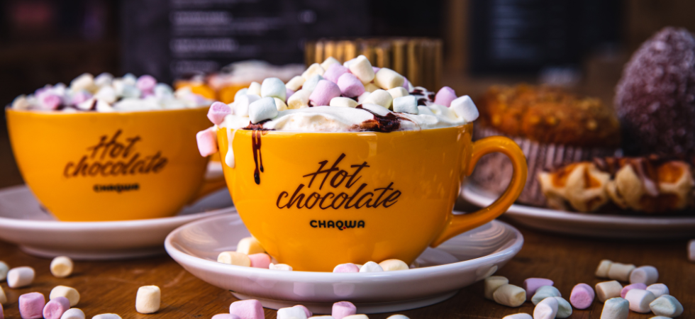 Två gula muggar märkta Hot chocolate CHAQUA fyllda med varm choklad, vispgrädde och minimarshmallows står på fat omgivna av marshmallows, med bakverk i bakgrunden - perfekt för en mysig skiferie i Geilo.
