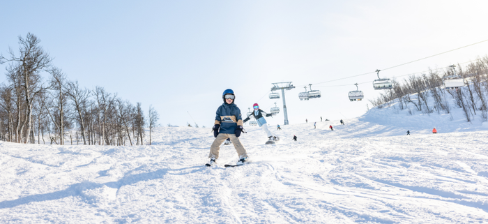 Ett barn i vinterkläder ler när det åker skidor nedför en snöig backe under en skiferie i Geilo, med andra skidåkare, lövträd och stolliftar synliga i den ljusa, soliga bakgrunden.