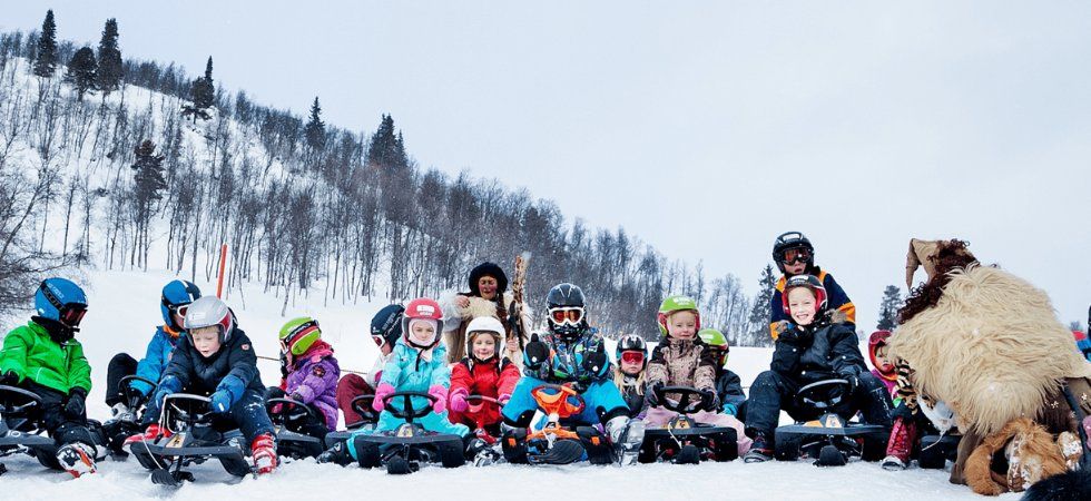 En grupp barn i vinterkläder och hjälmar sitter på slädar i snön och ler under sin skiferie i Geilo. I bakgrunden syns snötäckta träd och ett berg, bakom dem står två vuxna och en person i pälsdräkt.