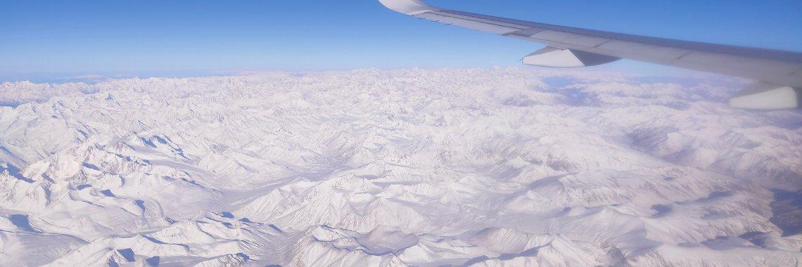 Flygfoto av en snötäckt bergskedja under en klarblå himmel, med en flygplansvinge synlig i övre högra hörnet - ett perfekt perspektiv för en skidsemester med flyg.