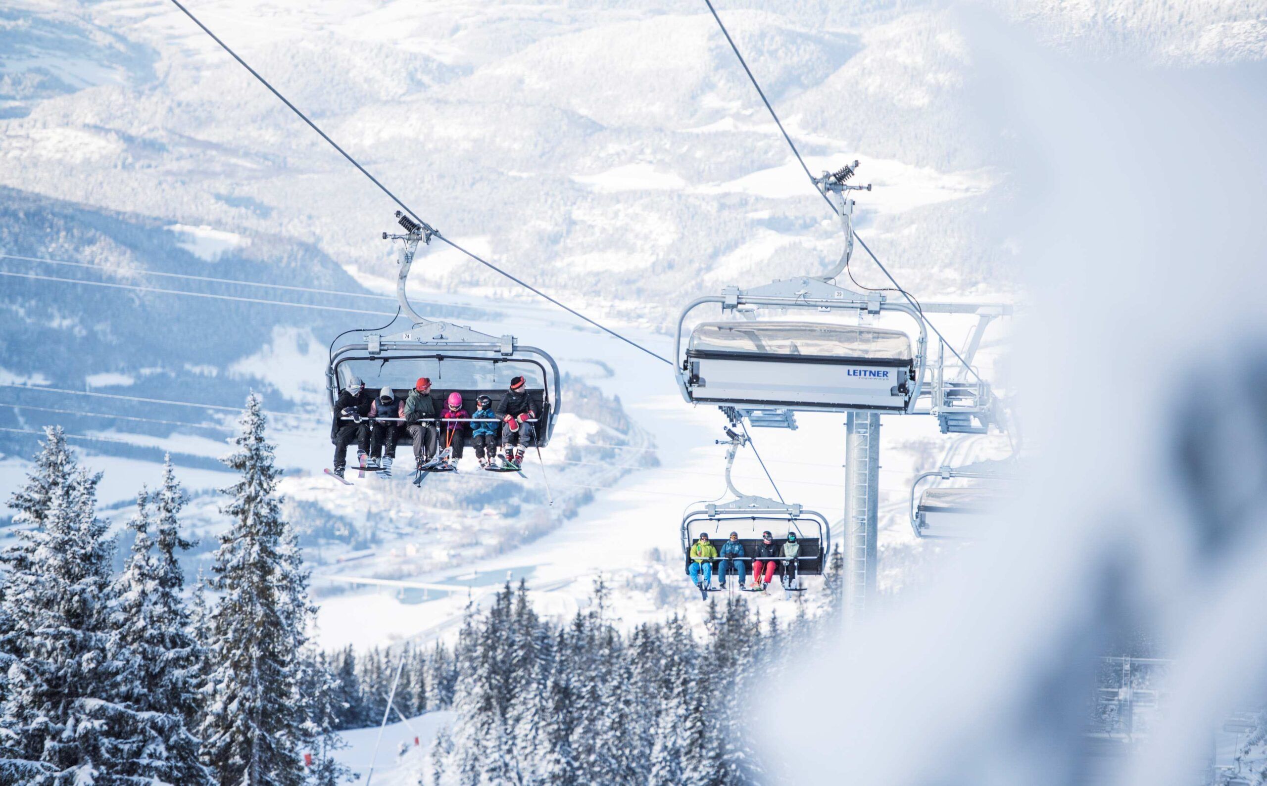 Et skisportssted udfolder sig, mens folk i stolelifte stiger op over Wagrains sneklædte landskab. Træer er støvet med sne, og fjerne bjerge danner baggrunden. Den klare himmel forstærker den vinterlige, fredelige atmosfære, og inviterer skirejseentusiaster til at fordybe sig i dette alpine paradis.