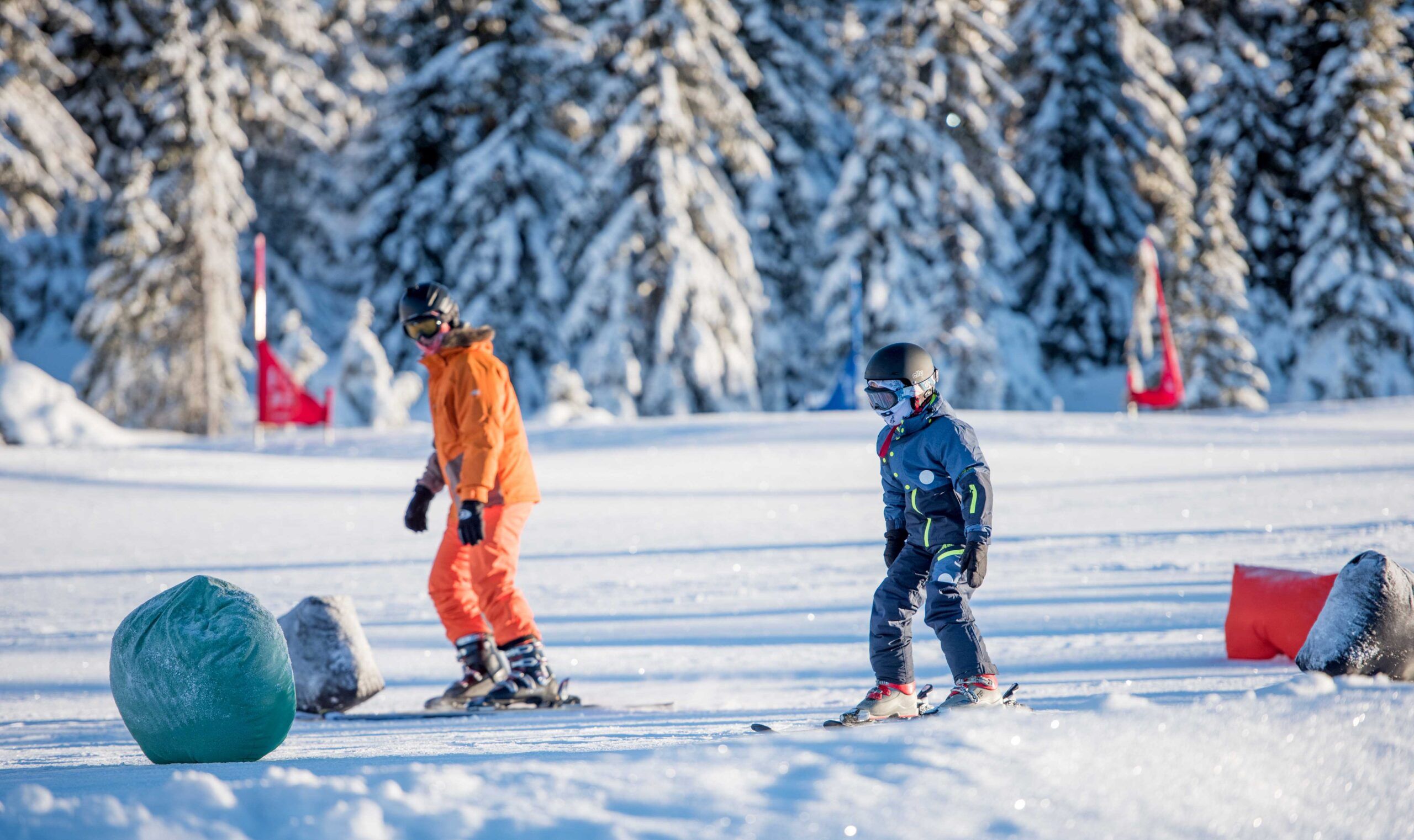To personer står på ski på en sneklædt skråning i Saalbach, omgivet af snedækkede træer. Den ene bærer et orange outfit og den anden et mørkt. Begge har hjelme og beskyttelsesbriller, med farverige flag i baggrunden, der fanger den spændende essens af skirejser.