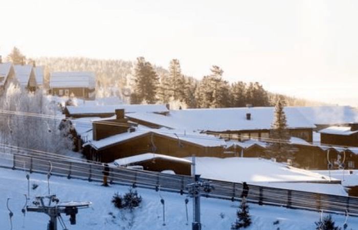 En snöig skidortsscen på Skiferie i Idre Fjäll med trästugor och byggnader omgivna av snötäckta träd. Skidliftsutrustningen syns i förgrunden under mjukt, starkt solljus.