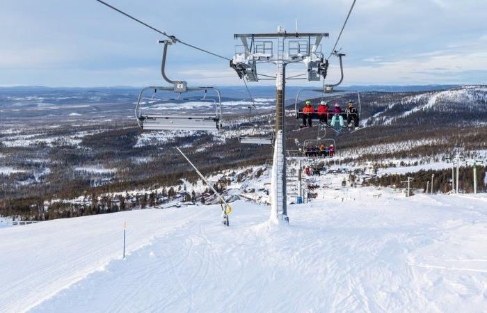 Människor åker skidlift uppför en snöig bergssluttning, med utsikt över träd och avlägsna kullar under en delvis molnig himmel. Detta vintersportområde fångar essensen av en skiferie i Idre Fjäll, med skidåkare som glider nedanför.