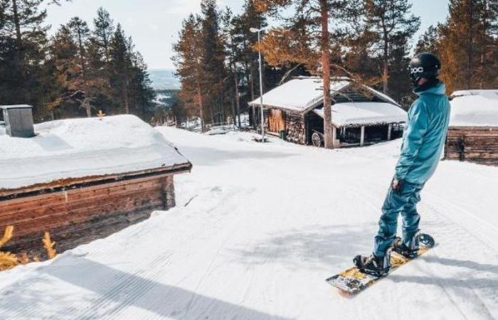 En snowboardåkare i blåkläder står på en snöig sluttning nära trästugor, omgiven av tallar under en klar himmel - det perfekta skiferie i Idre Fjäll.