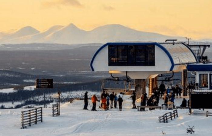 Människor samlas vid en skidliftstation på ett snöigt berg i solnedgången under sin skiferie i Idre Fjäll, med avlägsna bergstoppar synliga under en gyllene himmel.
