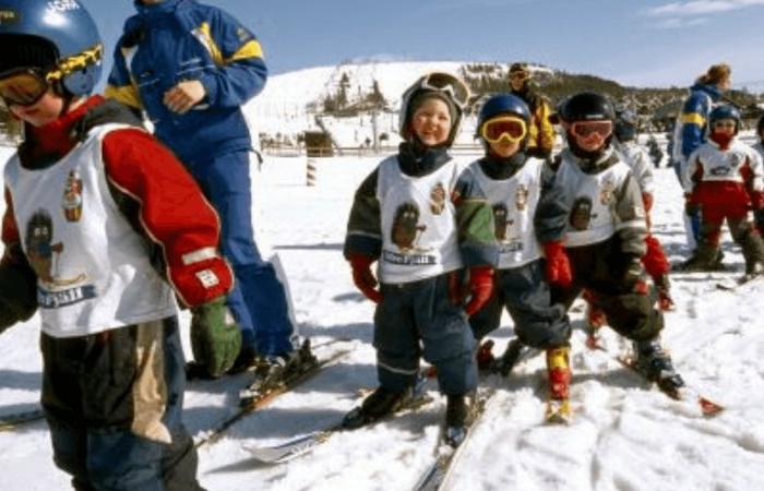 En grupp små barn i skidkläder, hjälmar och glasögon står i en rad i en snöig backe på Skiferie i Idre Fjäll, tillsammans med vuxna i blåställ, och förbereder sig för en skidlektion en solig vinterdag.