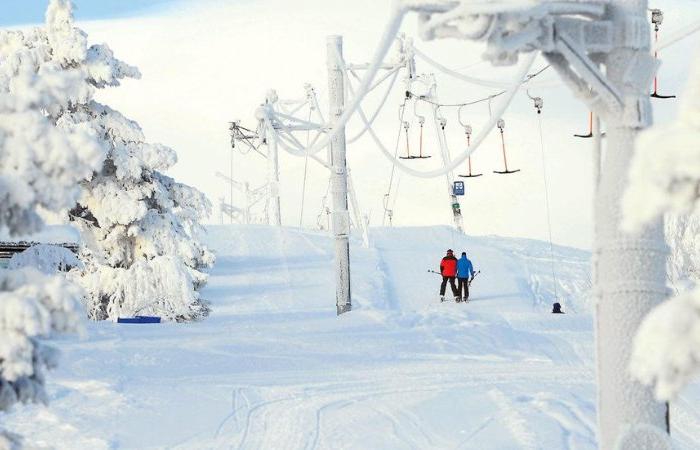 En snöig skidbacke med snötäckta träd, skidliftar och två personer i vinterkläder som står tillsammans nära liften en ljus och klar dag - perfekt för en skiferie i Idre Fjäll.