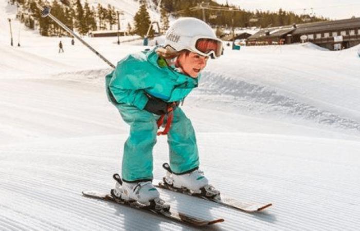 Ett litet barn i turkos skiddräkt, vit hjälm och skidglasögon åker utför i en snöig backe under en skiferie i Idre Fjäll, framåtböjd i skidställning med byggnader och träd i bakgrunden.