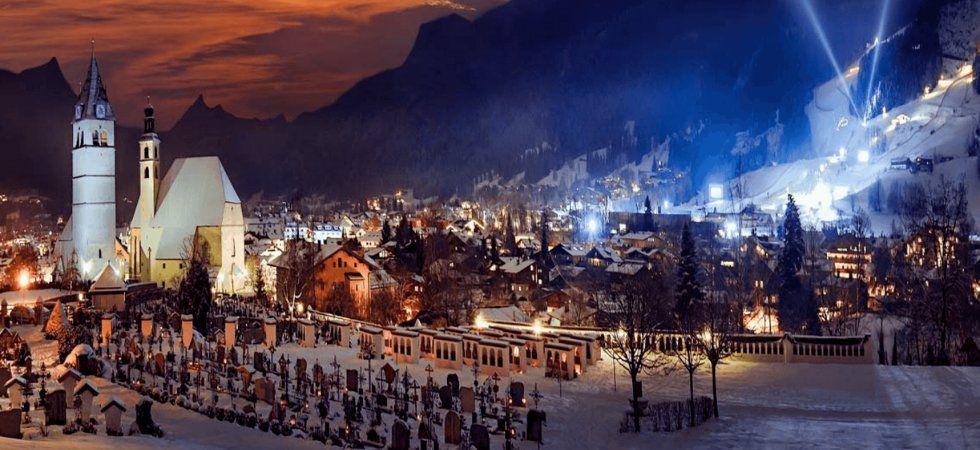 En snöig alpstad i skymningen, upplysta kyrkobyggnader och en kyrkogård i förgrunden, med starka lampor som lyser på en skidbacke - Skiferie i Kitzbühel erbjuder varma och kalla lampor som kontrasterar vackert mot bergsbakgrunden.