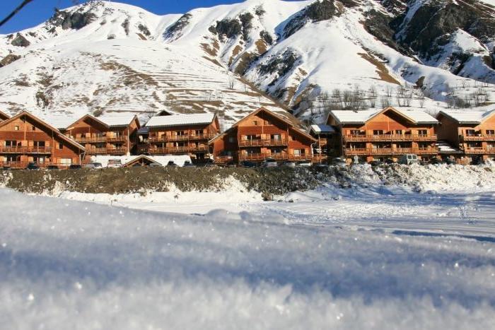 Snötäckta trästugor står på rad vid foten av karga, snöklädda berg under en klarblå himmel. Snön täcker marken i förgrunden och skapar en fridfull vinterscen.