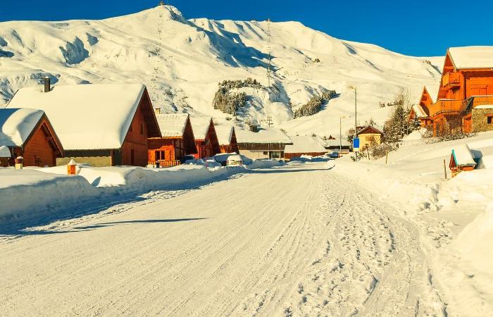 En snöig vinterby med trähus och snötäckta tak ligger vid foten av solbelysta berg under en klarblå himmel - den perfekta miljön för en skiferie i La Toussuire.
