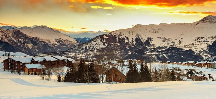 Snötäckt bergsby i solnedgången, med trästugor inbäddade bland träd och dramatiska, glödande orange moln över avlägsna snöiga toppar - perfekt för en skiferie i La Toussuire.