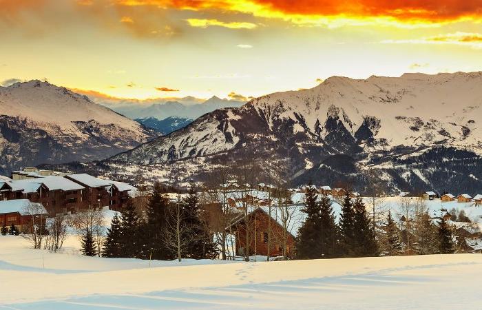 Snötäckt bergsby i solnedgången, med trästugor inbäddade bland träd och dramatiska, glödande orange moln över avlägsna snöiga toppar - perfekt för en skiferie i La Toussuire.