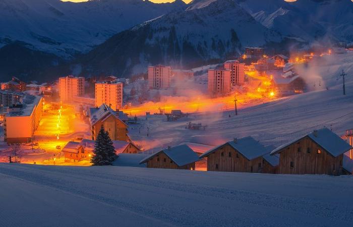 Snötäckt skidort inbäddad i bergen i skymningen, med varma ljussken från byggnader och gator - perfekt för en skiferie i La Toussuire - omgiven av snöiga sluttningar och mörka, skuggiga toppar i bakgrunden.