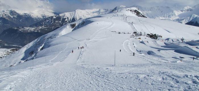 Ett snöigt bergslandskap med skidbackar, utspridda skidåkare och stolliftar sätter scenen för en perfekt skiferie i La Toussuire. Snötäckta toppar och avlägsna berg fyller bakgrunden under en molnig himmel.