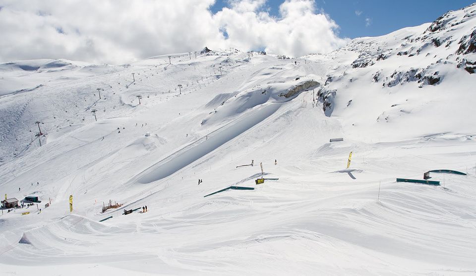 En snötäckt skidort i Wagrain har flera skidbackar, hinderbanor i terrängparker, rails och stolsliftar under en delvis molnig himmel, med några få skidåkare och snowboardåkare synliga i backarna.