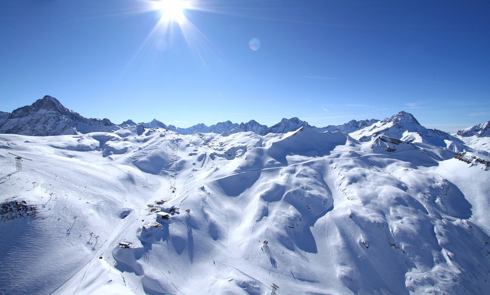 De snötäckta bergen i Wagrain sträcker sig under en klarblå himmel med solen som skiner starkt över huvudet. Landskapet har släta sluttningar, oländiga toppar och en fridfull, vinterlig atmosfär perfekt för skidresorsentusiaster.