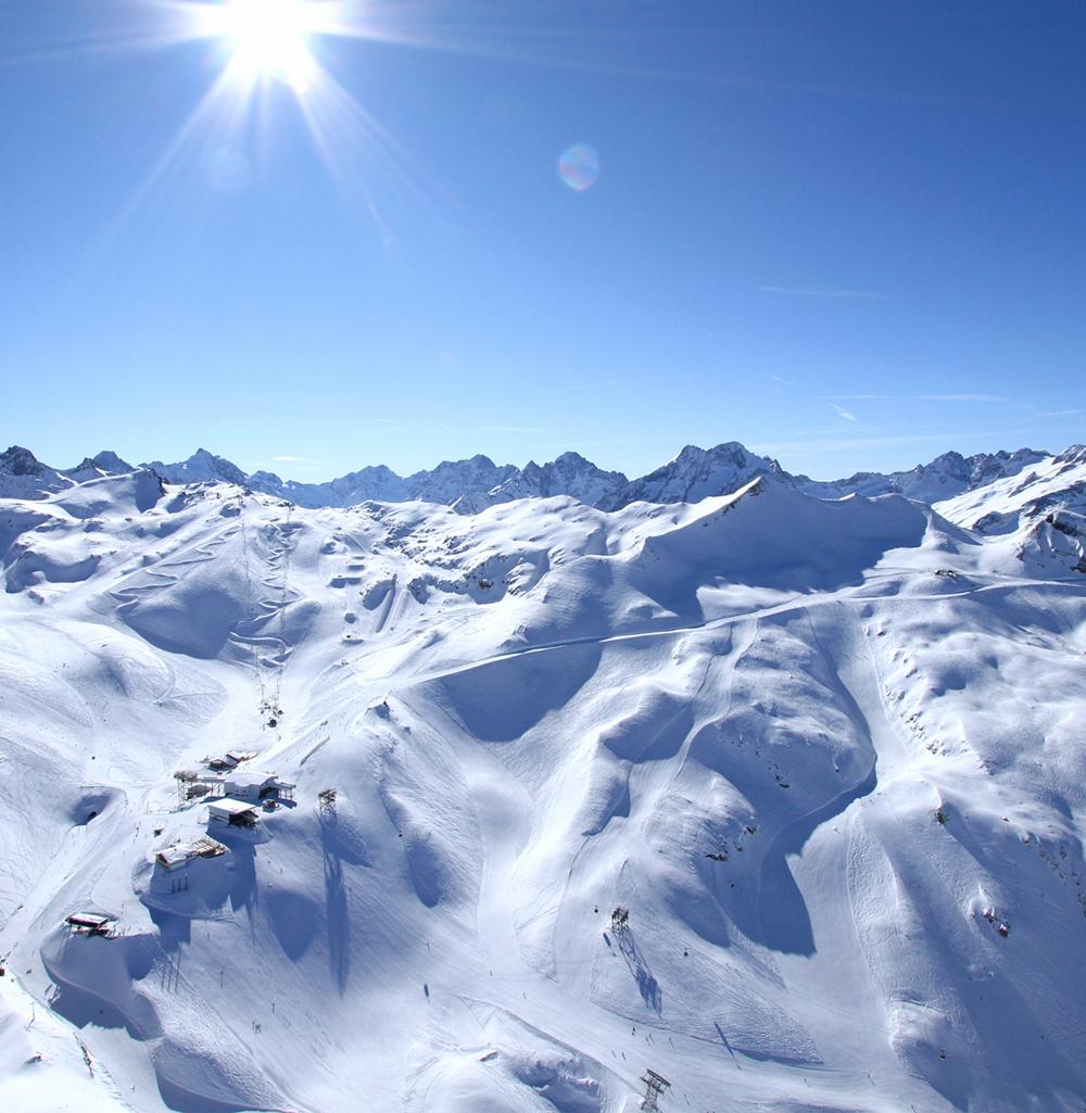 De snötäckta bergen i Wagrain sträcker sig under en klarblå himmel med solen som skiner starkt över huvudet. Landskapet har släta sluttningar, oländiga toppar och en fridfull, vinterlig atmosfär perfekt för skidresorsentusiaster.