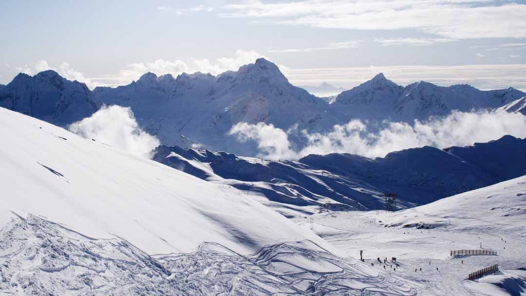 Snötäckta berg under en delvis molnig himmel, med skidspår och några skidåkare synliga i backarna. Spetsiga toppar reser sig i bakgrunden nära Westendorf, medan låga moln dröjer sig kvar bland bergen.