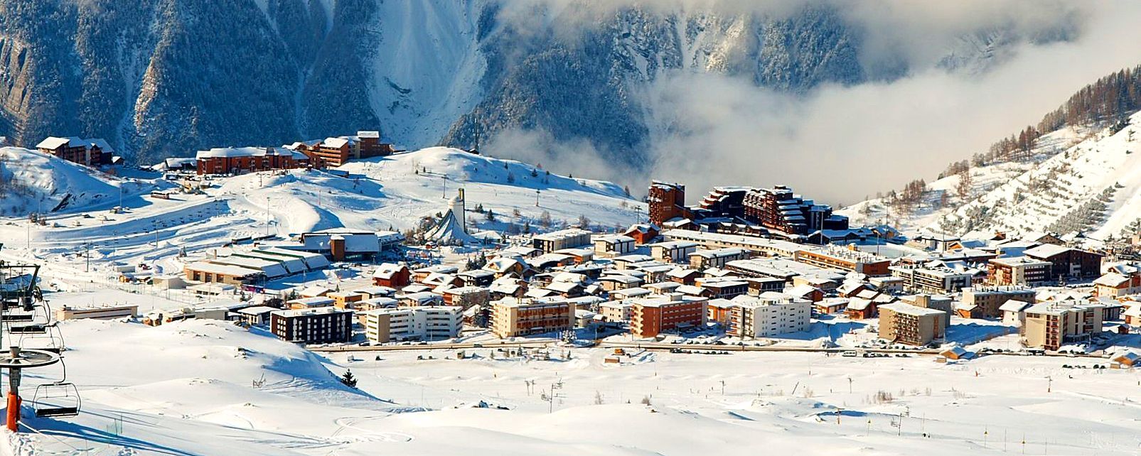 En snöig bergsby som Wagrain, med många byggnader och skidstugor inbäddade bland snötäckta kullar och träd, omgivna av berg och dimma.