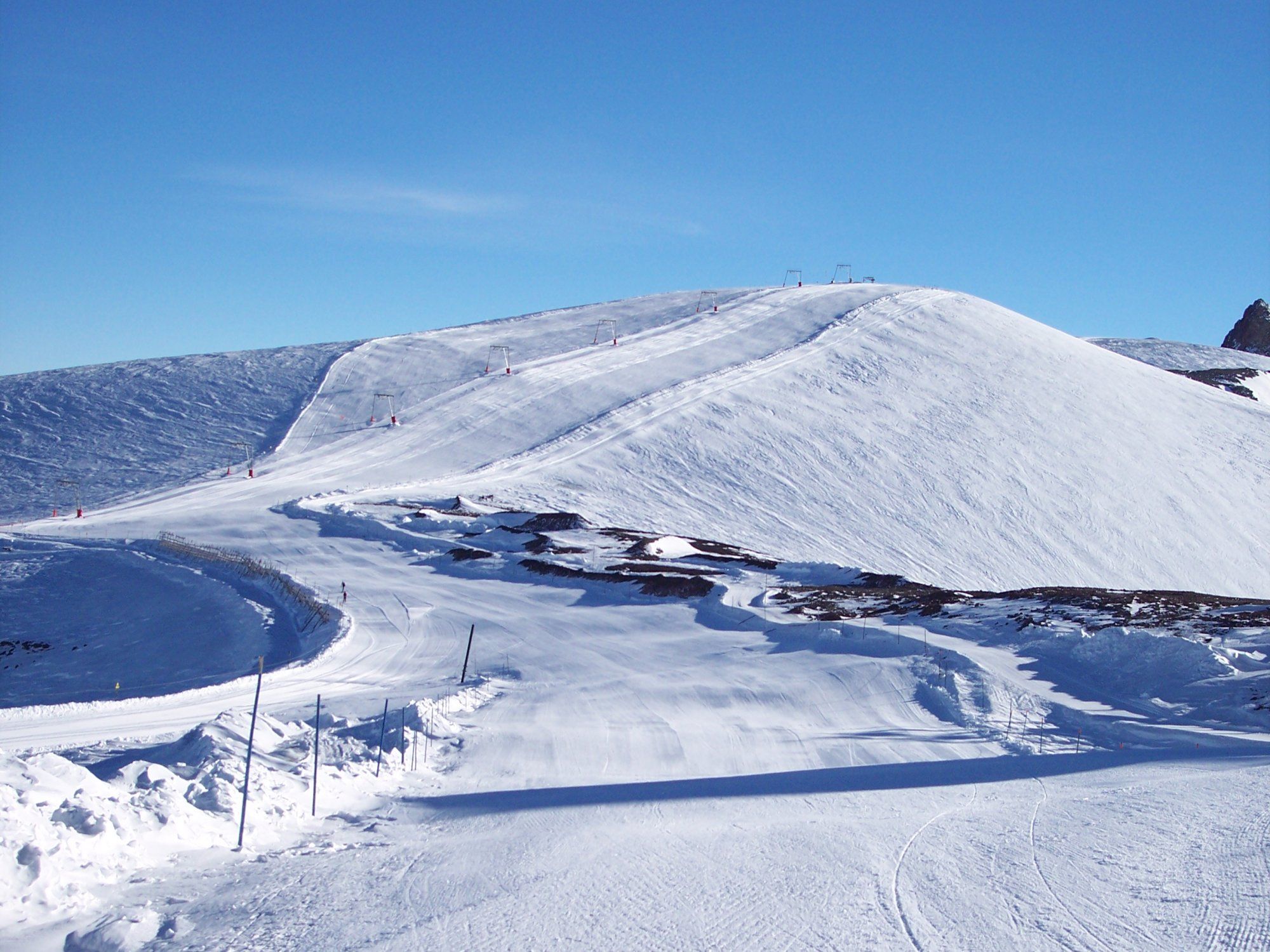 En snötäckt skidbacke i Saalbach, under klarblå himmel. Spår korsar snön och skidmarkörer kantar de fridfulla stigarna. Det orörda landskapet utspelar sig med mjuka sluttningar som leder upp till en liten kulle.