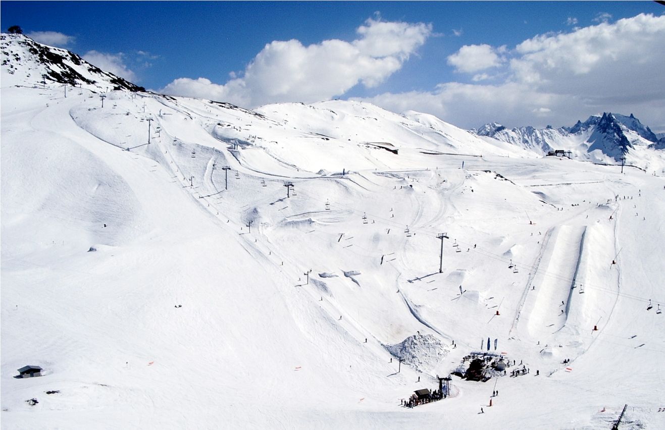 En snörik skidort som Saalbach, med flera skidbackar, skidliftar och några skidåkare utspridda över landskapet. Scenen utspelar sig mot en bakgrund av karga toppar och delvis molnig himmel.