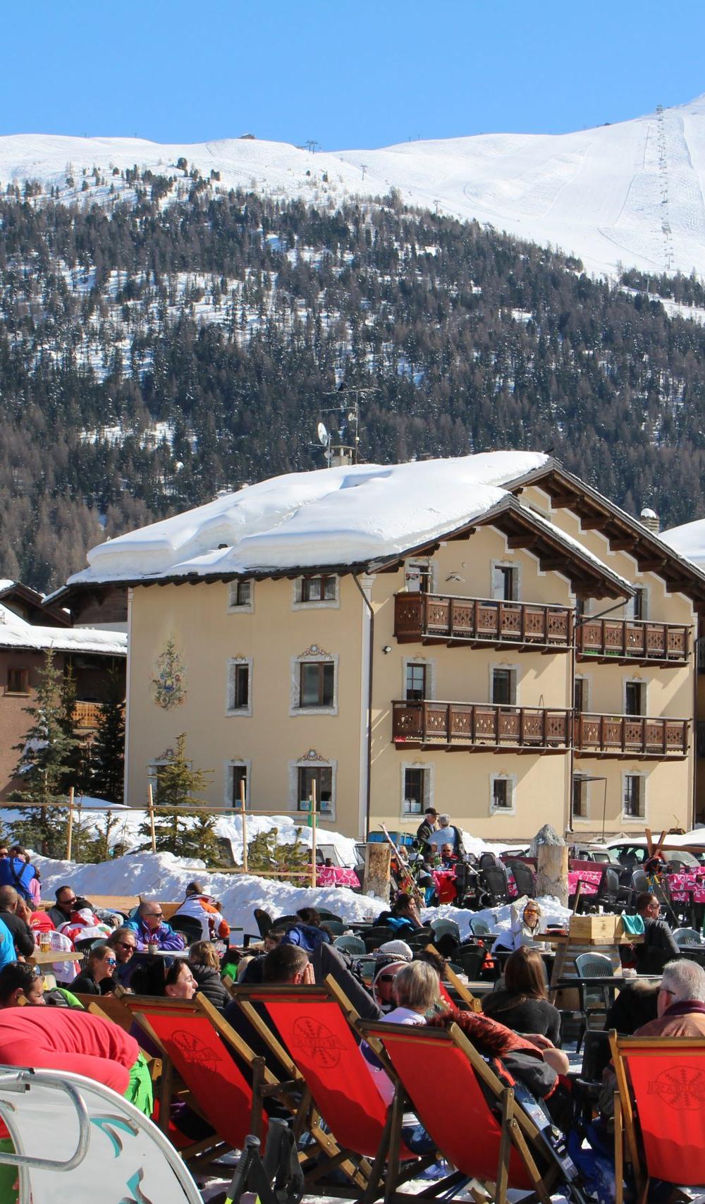 Et travlt skisportssted i Westendorf byder på folk, der slapper af i liggestole og nyder skiferiestemningen. Snedækkede bjerge og bygninger i chalet-stil pryder baggrunden under en klar blå himmel, mens ski og snowboards er støttet i nærheden.