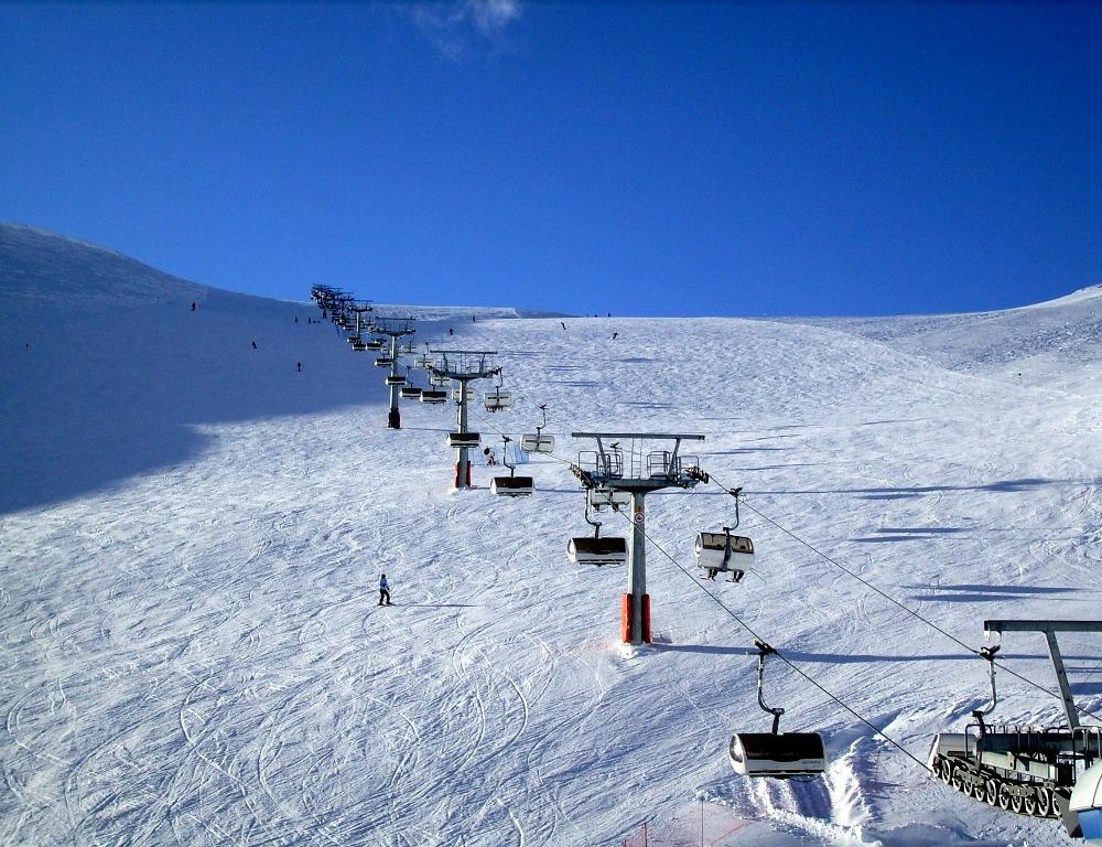 En snöig skidbacke i Saalbach med en skidlift uppför centrum. Några skidåkare navigerar nerför backen under en klarblå himmel. Liften har flera stolar, och spår från tidigare skidåkare kan ses i snön, vilket gör den till en perfekt plats för en minnesvärd skidsemester.