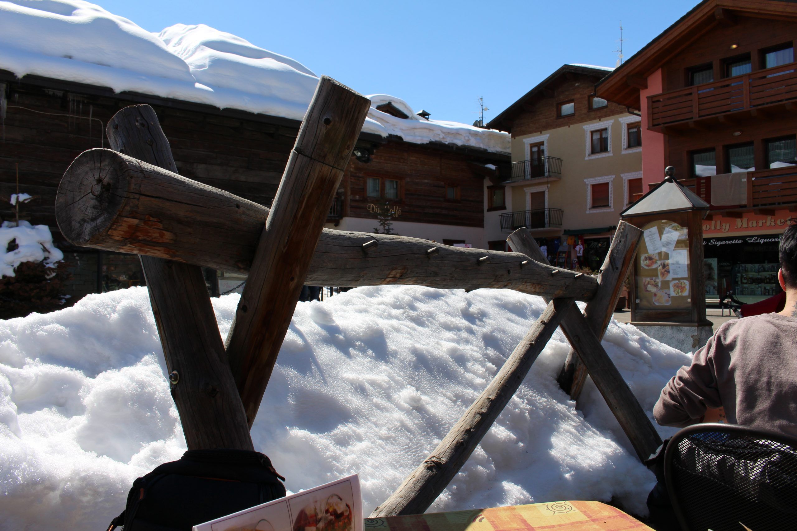 En sneklædt scene i en bjerglandsby, perfekt til en skiferie, har træhegn delvist dækket af sne. Træ- og stenbygninger med altaner står under en klar blå himmel. I forgrunden ses en del af et bord med en menu og en person i jakke, som fanger den hyggelige skistemning.