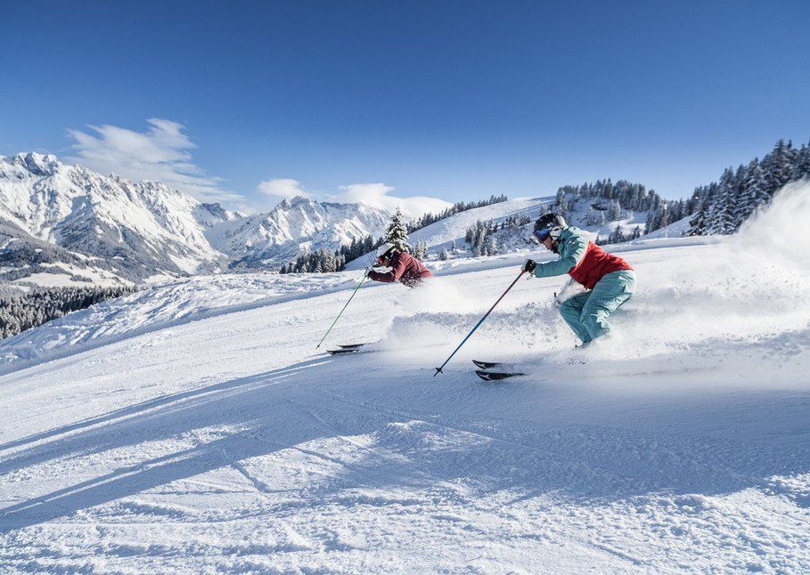 Två personer åker skidor utför på en snöig bergssluttning i Wagrain, omgivna av snötäckta träd och bergstoppar, under en klarblå himmel – en perfekt scen för oförglömliga skidresor.