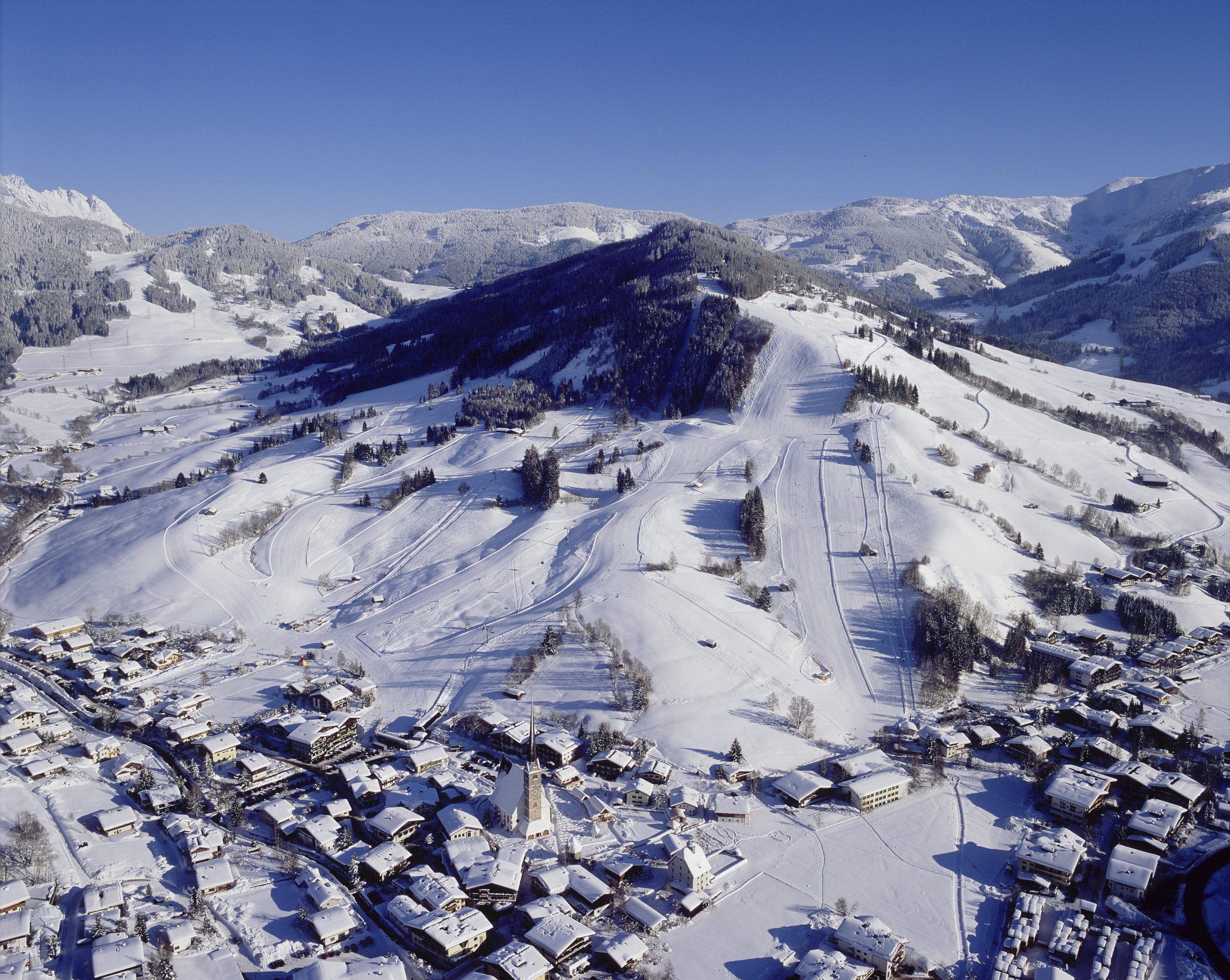 En snöig alpby ligger vid foten av en skidort i Wagrain, med trädkantade backar och slingrande skidspår, omgiven av snötäckta berg under en klarblå himmel – perfekt för en minnesvärd skidresa.