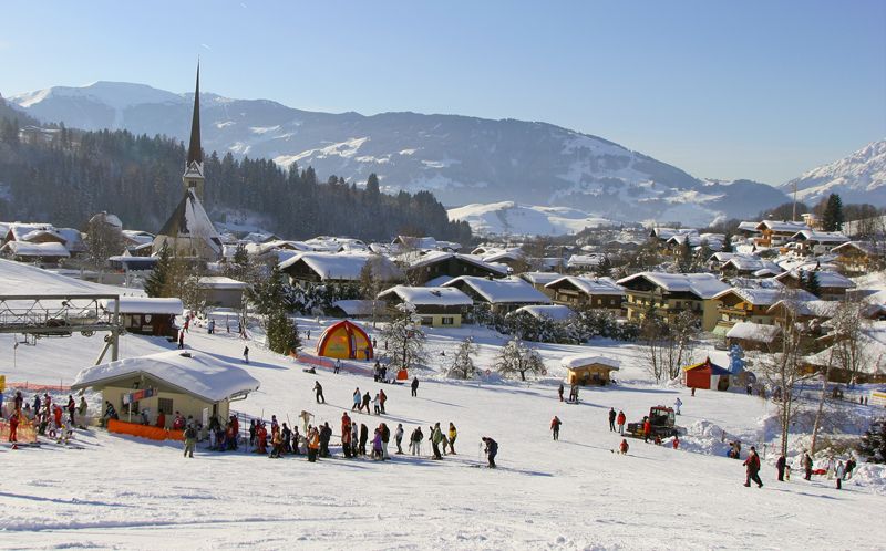 En snöig skidortsby i Saalbach med människor som åker skidor och samlas nära skidliftar, alphus utspridda över landskapet, en hög kyrktorn och skogsklädda berg – perfekt för din nästa skidresa eller skidresort under en klar himmel.