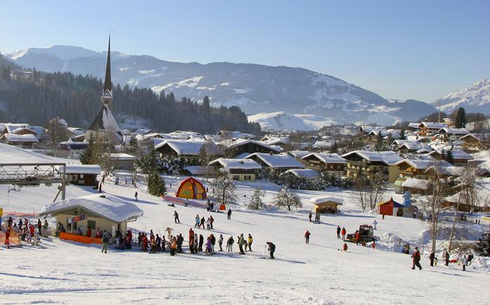 En snöig skidortsby i Saalbach med människor som åker skidor och samlas nära skidliftar, alphus utspridda över landskapet, en hög kyrktorn och skogsklädda berg – perfekt för din nästa skidresa eller skidresort under en klar himmel.