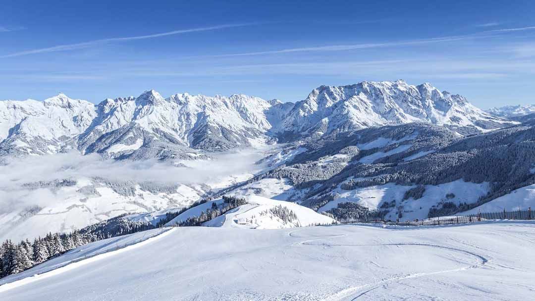 Snötäckta berg under en klarblå himmel, med spridda moln och skogar på sluttningarna. Böljande kullar och dalar är täckta av snö, vilket skapar ett lugnt vinterlandskap – perfekt för skidåkning och minnesvärda skidresor.
