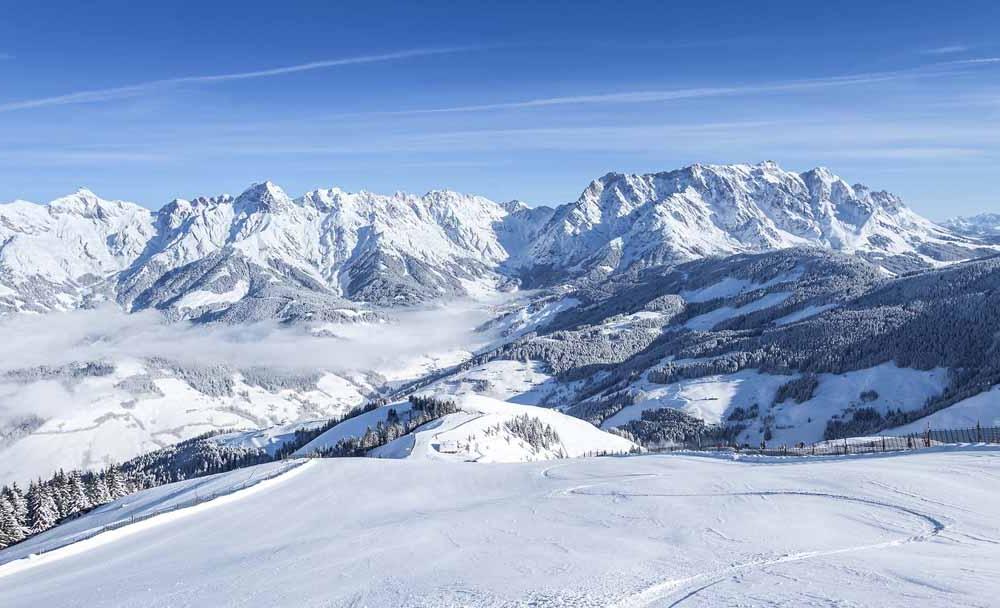 Snötäckta berg under en klarblå himmel, med spridda moln och skogar på sluttningarna. Böljande kullar och dalar är täckta av snö, vilket skapar ett lugnt vinterlandskap – perfekt för skidåkning och minnesvärda skidresor.