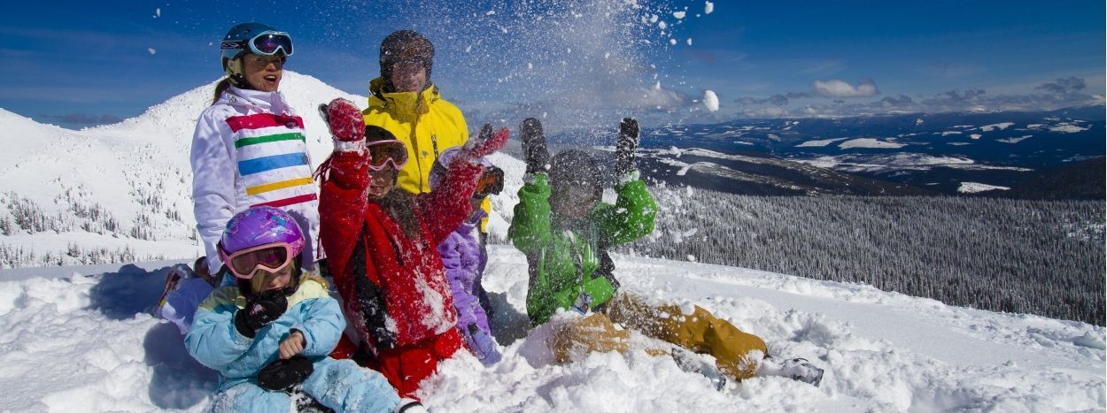 En grupp människor klädda i färgglada vinterkläder leker och skrattar tillsammans i djup snö på ett soligt berg, med snötäckta träd och avlägsna kullar i bakgrunden.