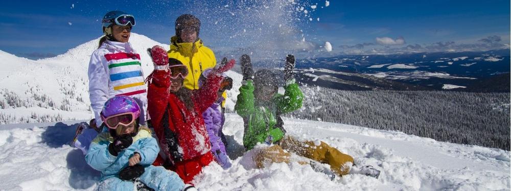 En grupp människor klädda i färgglada vinterkläder leker och skrattar tillsammans i djup snö på ett soligt berg, med snötäckta träd och avlägsna kullar i bakgrunden.