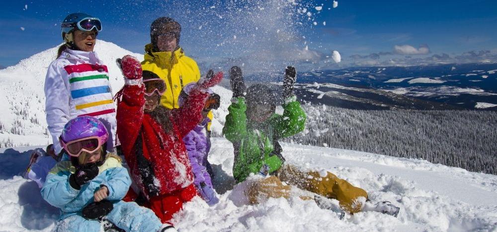 En grupp människor klädda i färgglada vinterkläder leker och skrattar tillsammans i djup snö på ett soligt berg, med snötäckta träd och avlägsna kullar i bakgrunden.