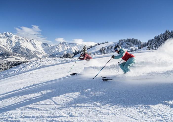 To skiløbere i farverigt gear suser ned ad en sneklædt bjergskråning i Maria Alm, med forrevne, snedækkede tinder og stedsegrønne træer under en klar blå himmel, hvilket giver den perfekte skiferie.
