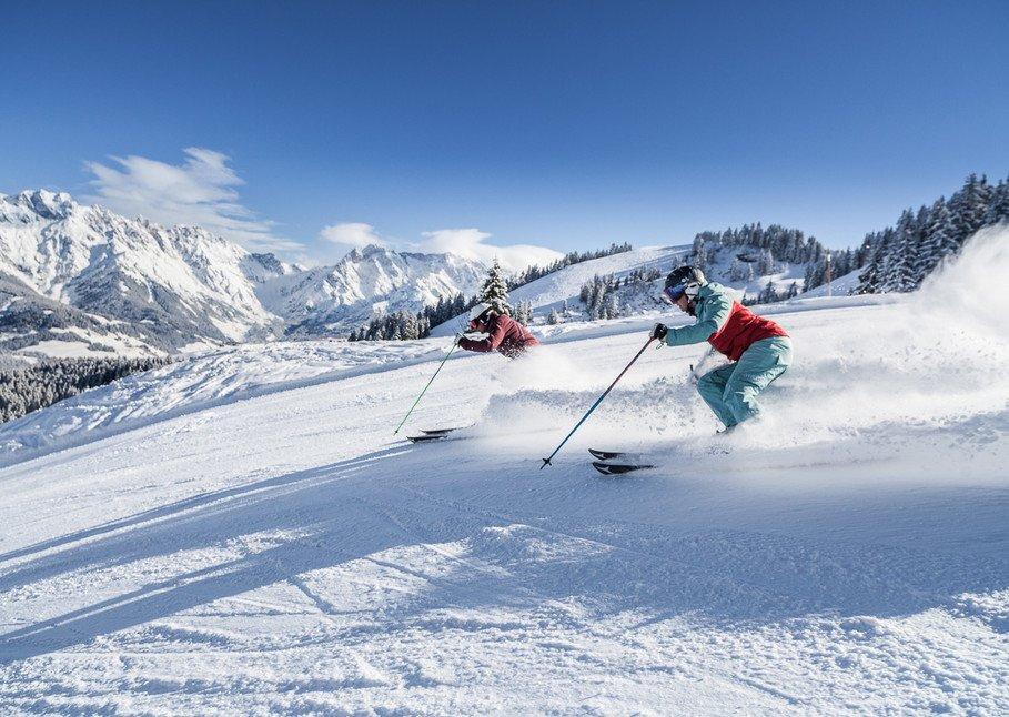 To skiløbere i farverigt gear suser ned ad en sneklædt bjergskråning i Maria Alm, med forrevne, snedækkede tinder og stedsegrønne træer under en klar blå himmel, hvilket giver den perfekte skiferie.