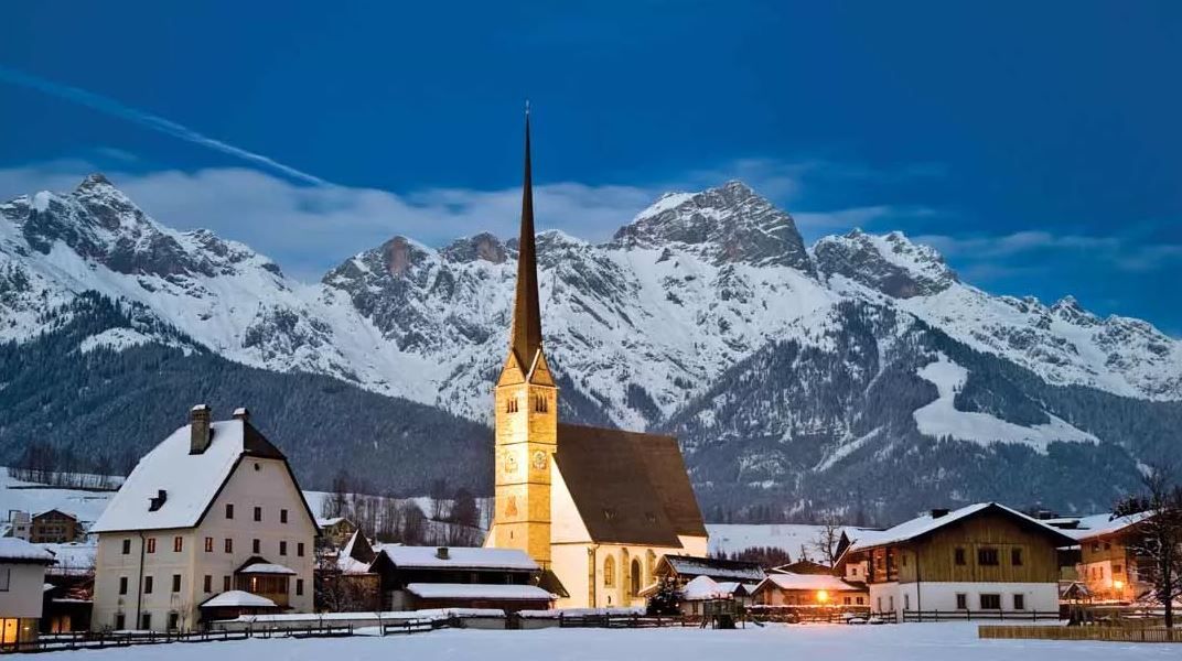 En snötäckt alpby i skymningen, med Westendorfs höga kyrktorn upplyst i mitten, omgiven av trähus och dramatiska, snötäckta berg i bakgrunden.