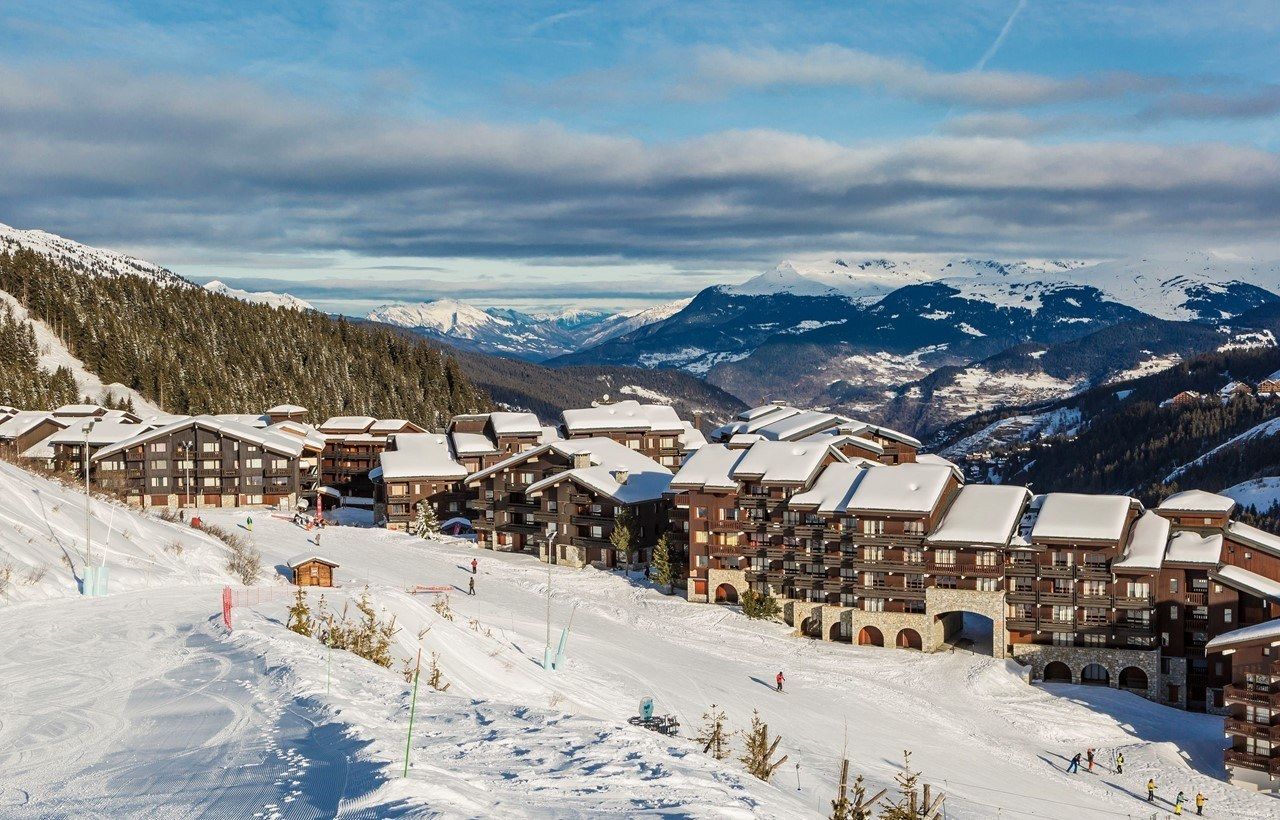 En snöig skidort med byggnader i chalet-stil som ligger inbäddad i bergen, omgiven av tallar och skidbackar, med skidåkare som syns och avlägsna snötäckta toppar under en delvis molnig himmel.