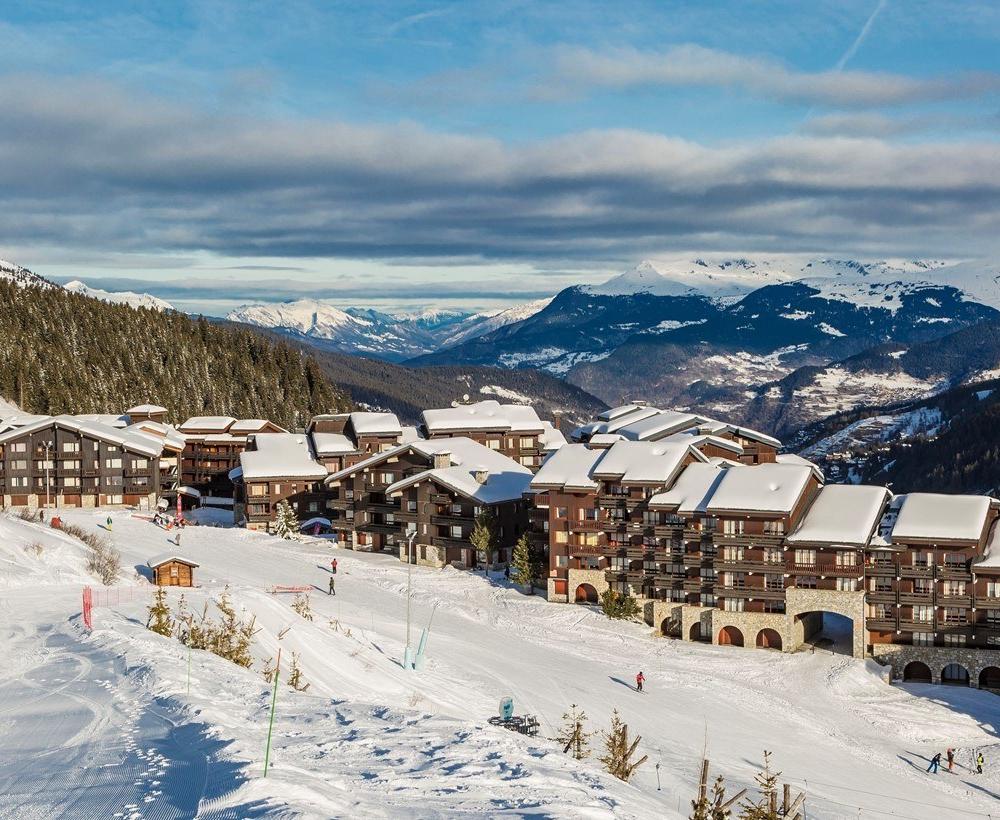 En snöig skidort med byggnader i chalet-stil som ligger inbäddad i bergen, omgiven av tallar och skidbackar, med skidåkare som syns och avlägsna snötäckta toppar under en delvis molnig himmel.