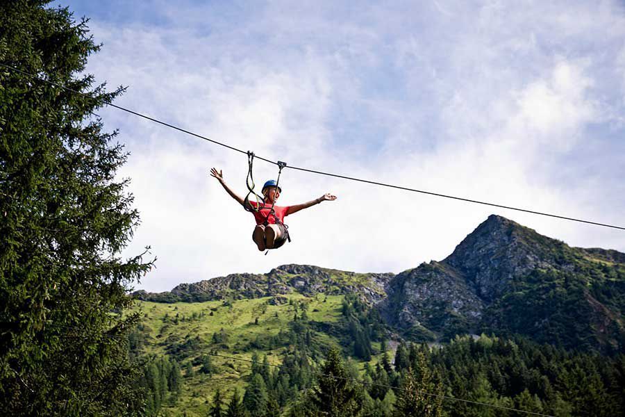 En person i en rød skjorte nyder at zipline med strakte arme, på baggrund af frodige grønne bjerge og en delvist overskyet himmel. Høje træer indrammer eventyret og fanger essensen af en spændende Sommerferie i Saalbach.