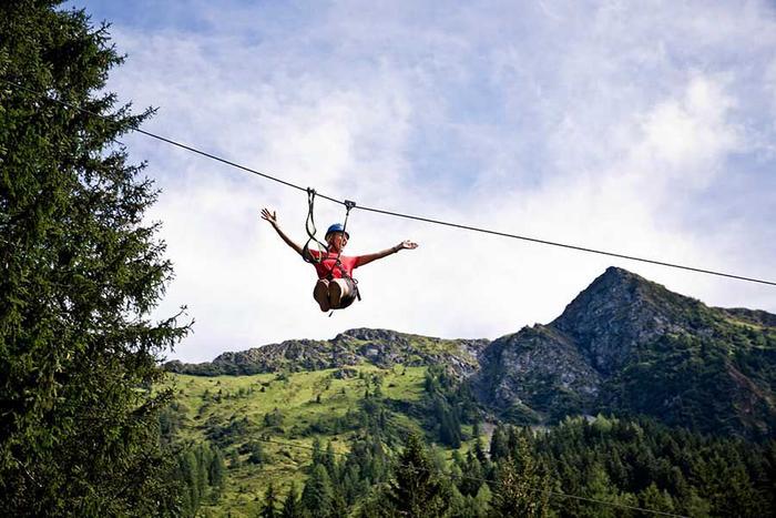 En person i en rød skjorte nyder at zipline med strakte arme, på baggrund af frodige grønne bjerge og en delvist overskyet himmel. Høje træer indrammer eventyret og fanger essensen af en spændende Sommerferie i Saalbach.