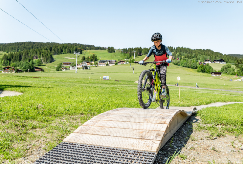 Et barn iført hjelm cykler over en lille trærampe i en græsmark og fanger eventyrlysten i et sommerferie Østrig. Baggrunden viser Saalbachs bakkede landskab med spredte huse og træer under en klar himmel.