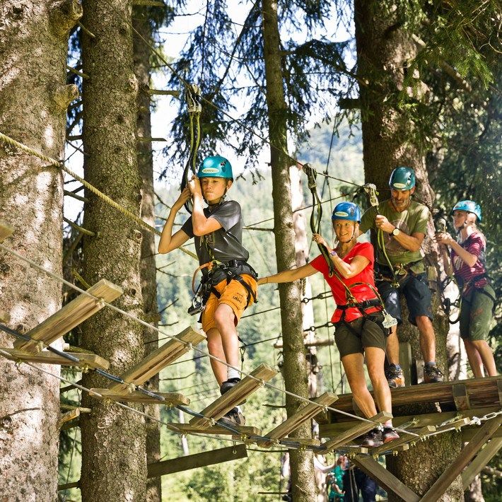 Under Sommerferie i Saalbach tager voksne og børn hjelme og seler på for at navigere på en trætop-rebbane, omgivet af høje træer og frodigt grønt på en solskinsdag.