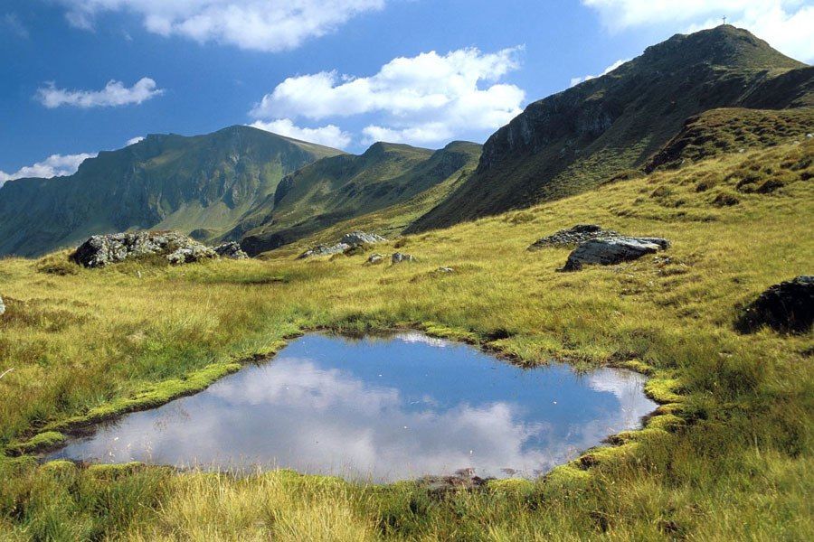 Et fredfyldt bjerglandskab, der minder om en sommerferie i Saalbach, har en lille reflekterende dam omgivet af græsklædt terræn. Klippede bjergskråninger rejser sig i baggrunden under en delvist overskyet blå himmel og indfanger roen i dette maleriske tilflugtssted.
