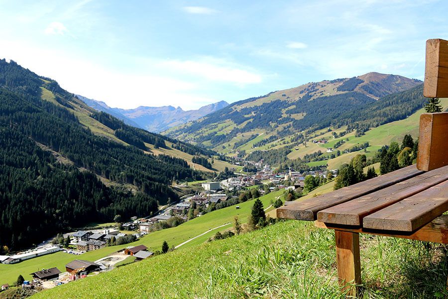 En naturskøn udsigt over en dal omgivet af grønne bakker og bjerge. Under en Sommerferie i Saalbach finder du en træbænk til højre, som tilbyder et malerisk sted med udsigt over landsbyen nedenfor under en klar blå himmel.